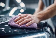 A man's hand wiping a wet car hood with a purple microfiber cloth, showcasing a car wash and detailing process.
